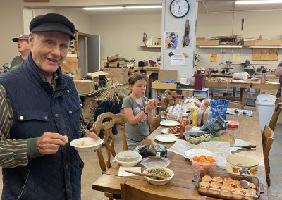 Man with bowl at potluck in midroom. Shared food is on the table.
